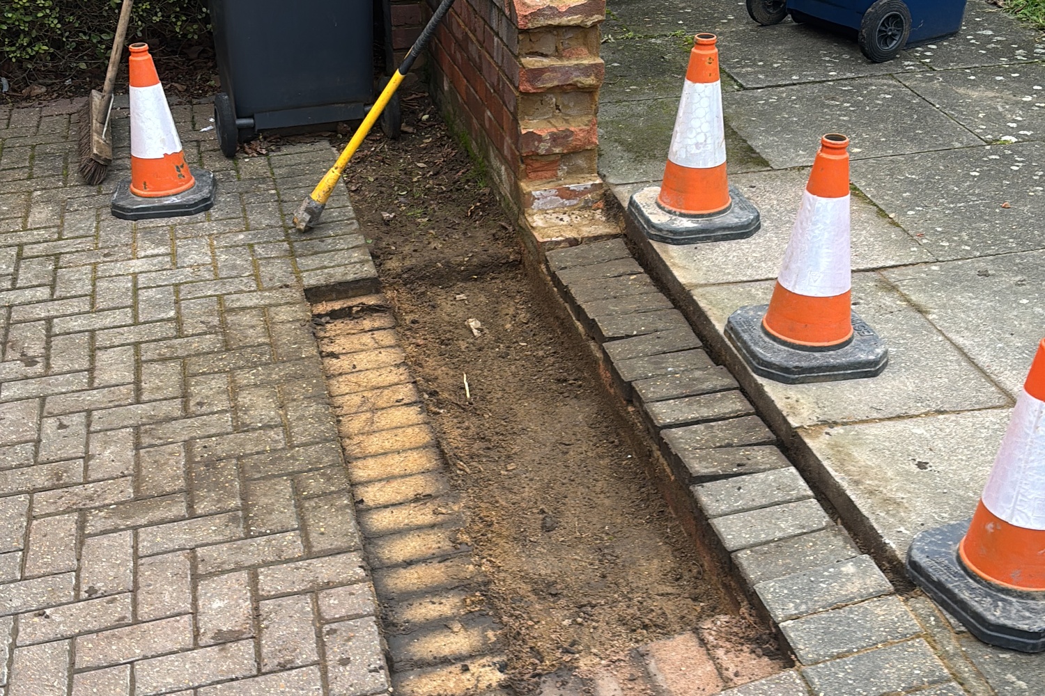 Open trench between block paving and brick wall with traffic cones — mid-job